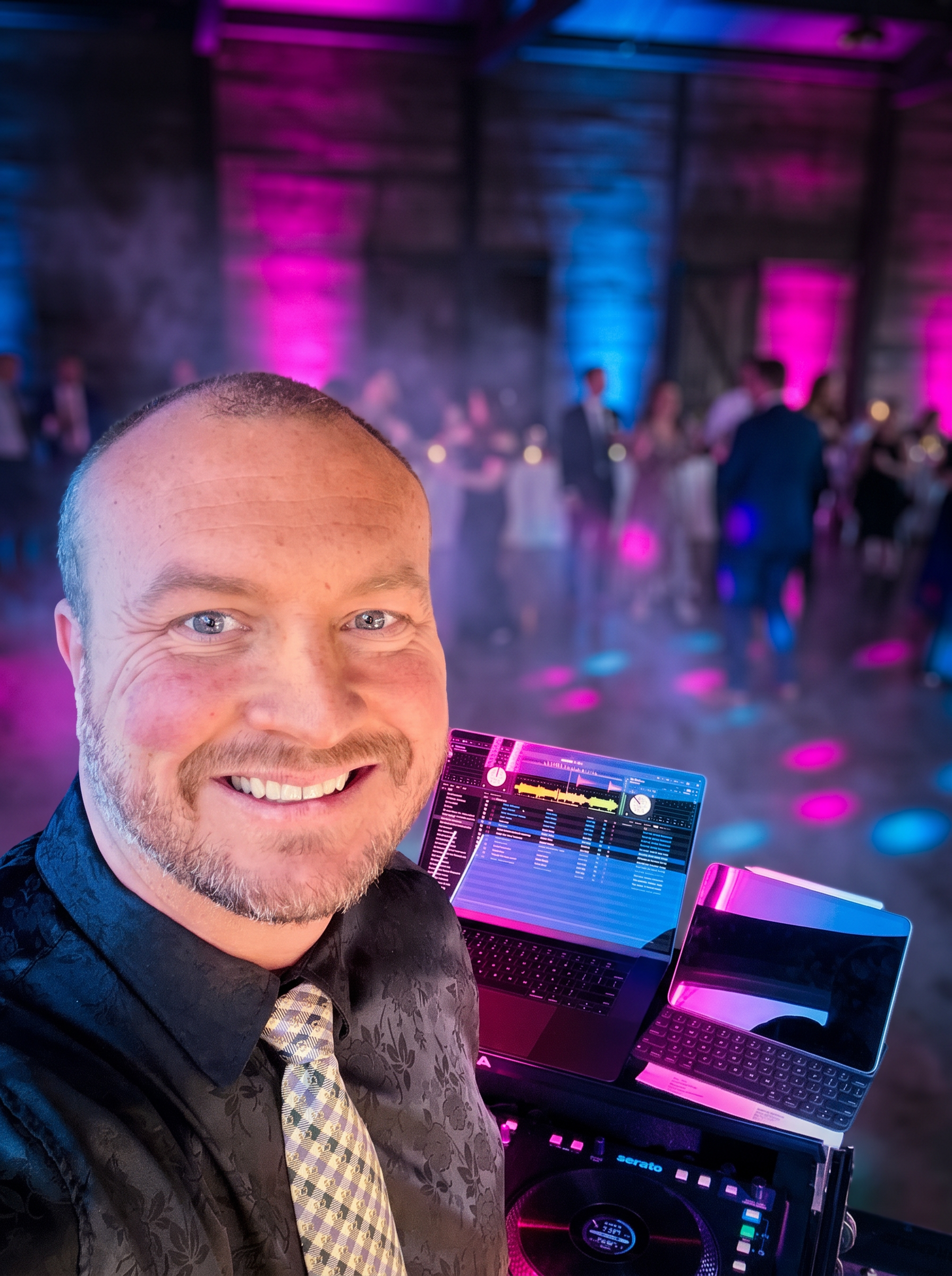 Craig Kraus, Portland wedding DJ, smiling behind his DJ setup with neon magenta and blue uplighting on the dance floor